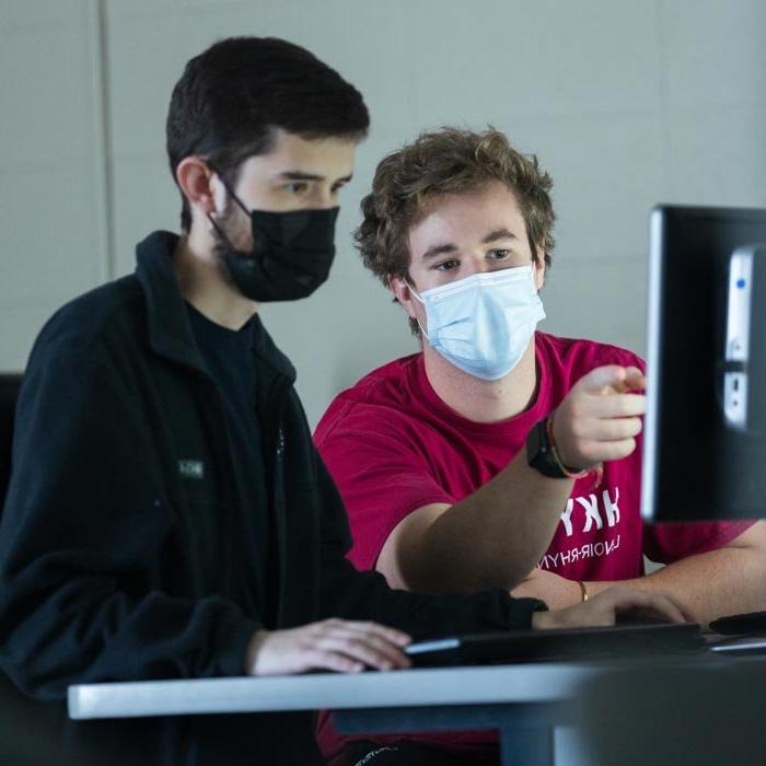 Two masked male students working on computer assignment in classroom